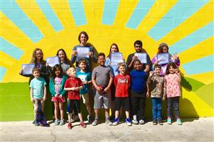 Photo of children and volunteers standing in front of a painted wall. The painting is a sun.